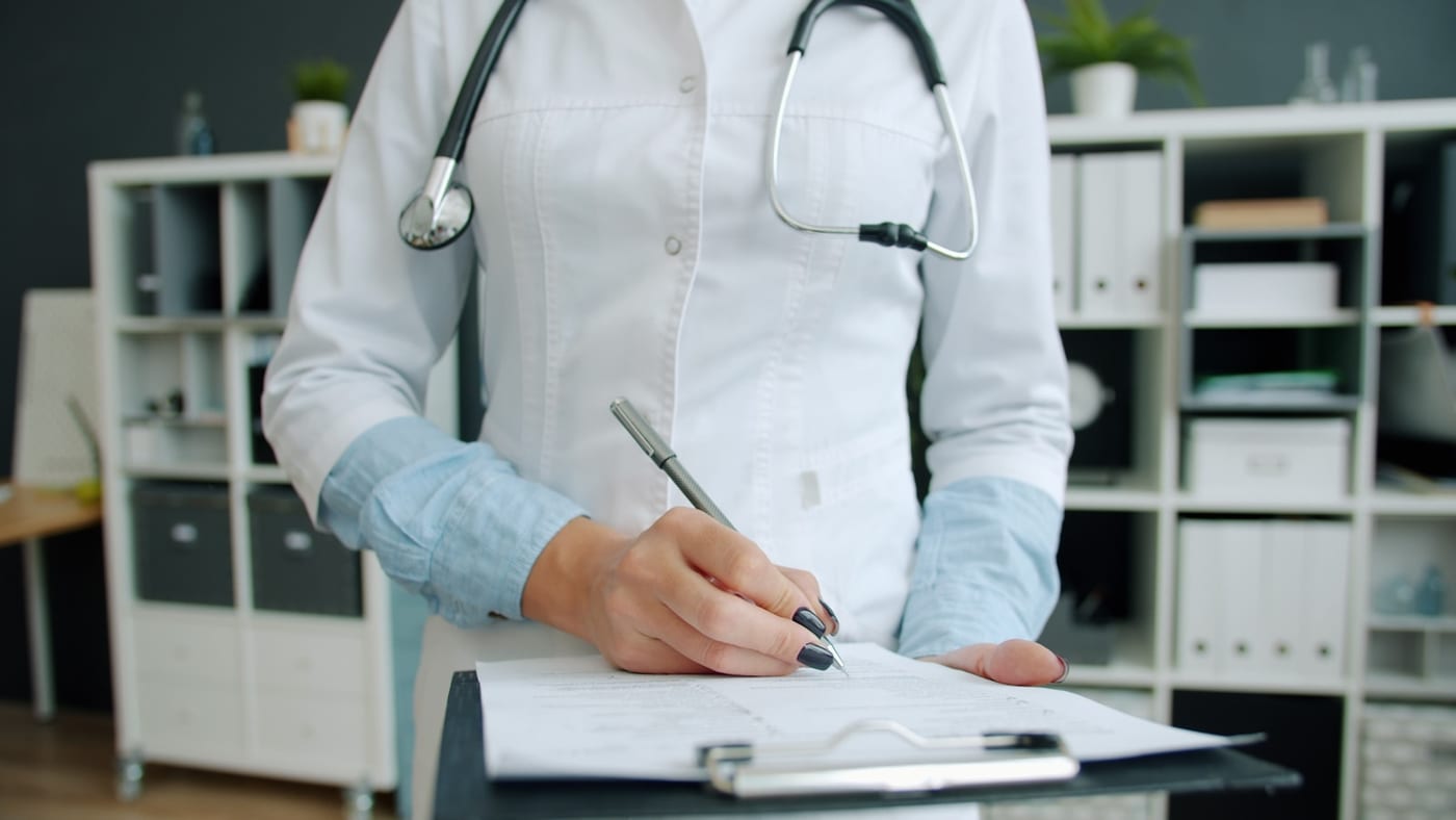 A clinician wearing a stethoscope writing notes on paper held on a clipboard, seen from the chest down.