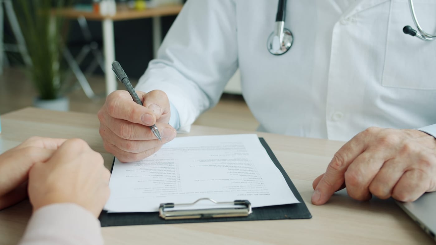 A clinician in a white coat writing on a printed form held on a clipboard at a desk, a patient's hands resting opposite.